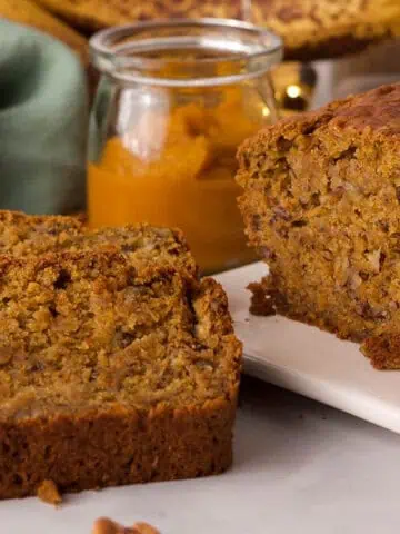 pumpkin banana bread loaf and slice propped next to it with ingredients in the background on counter with green tea towel
